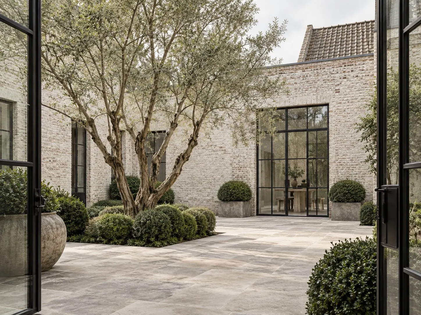 Quiet Belgian courtyard with limewashed masonry, black framed windows, limestone paving and sculptural trees.